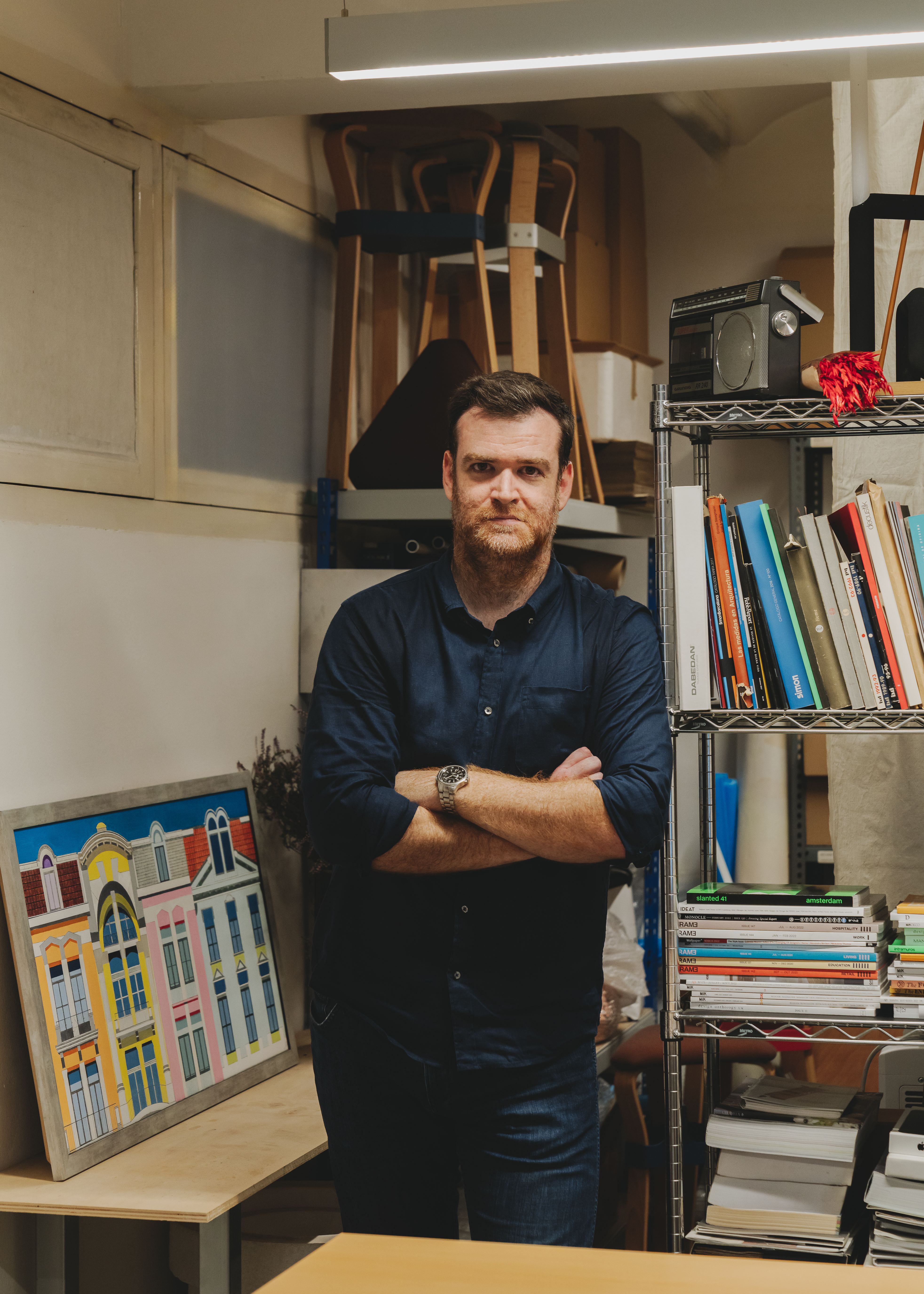 Portrait of designer Francesc Gasch standing in his studio beside a metal shelf filled with design books and objects. He wears a navy button-up shirt with arms crossed and a wristwatch visible. Behind him are stacked wooden stools, a vintage radio, and a colorful framed illustration of European-style buildings resting on a desk.