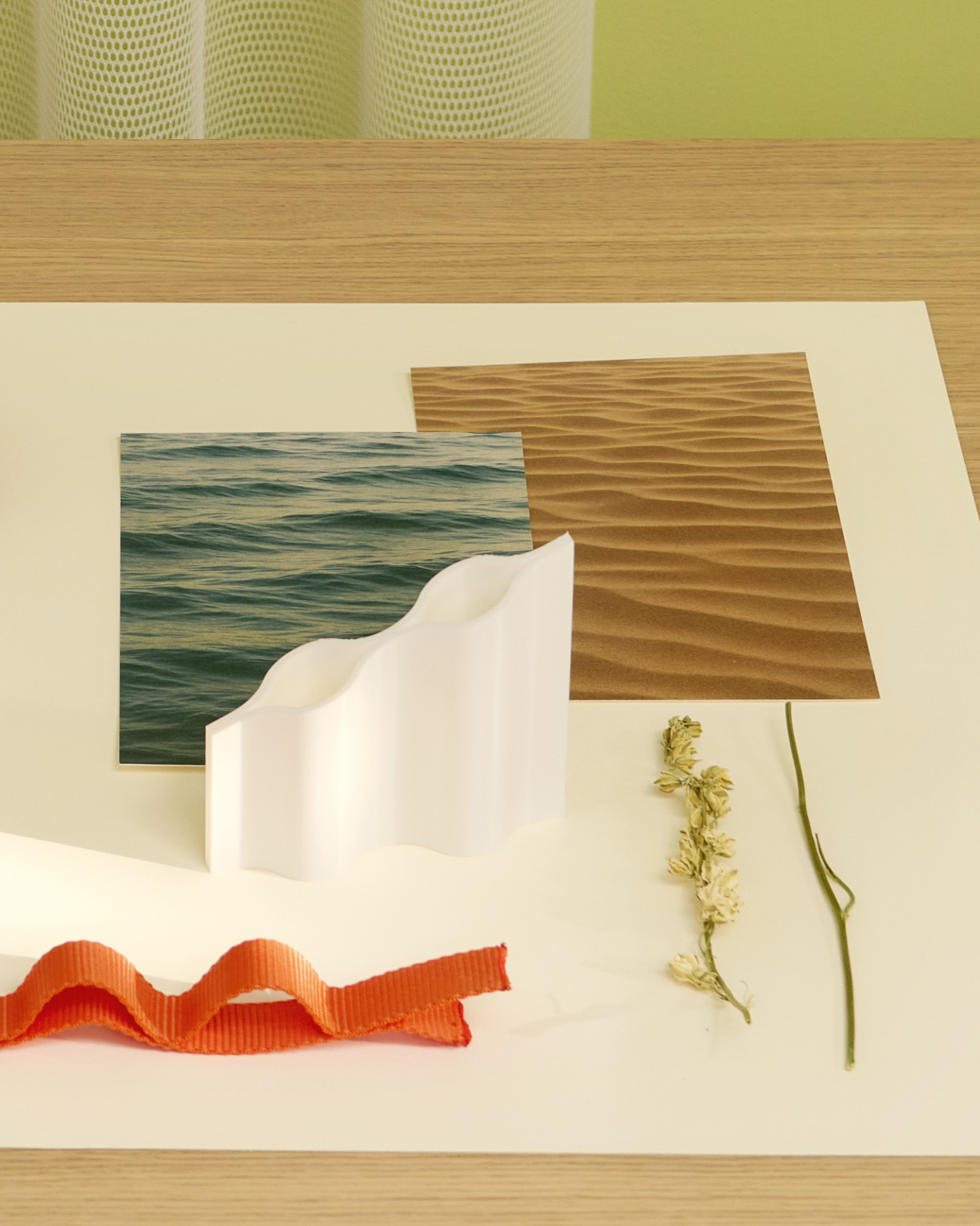 Inspiration board for the “Waves” vase by Francesc Gasch, featuring a white scale model, orange wave-shaped ribbon, dried botanical samples, and photos of ocean ripples and desert sand dunes. The elements are arranged on a table in front of a light green mesh curtain, echoing rhythmic natural flows.