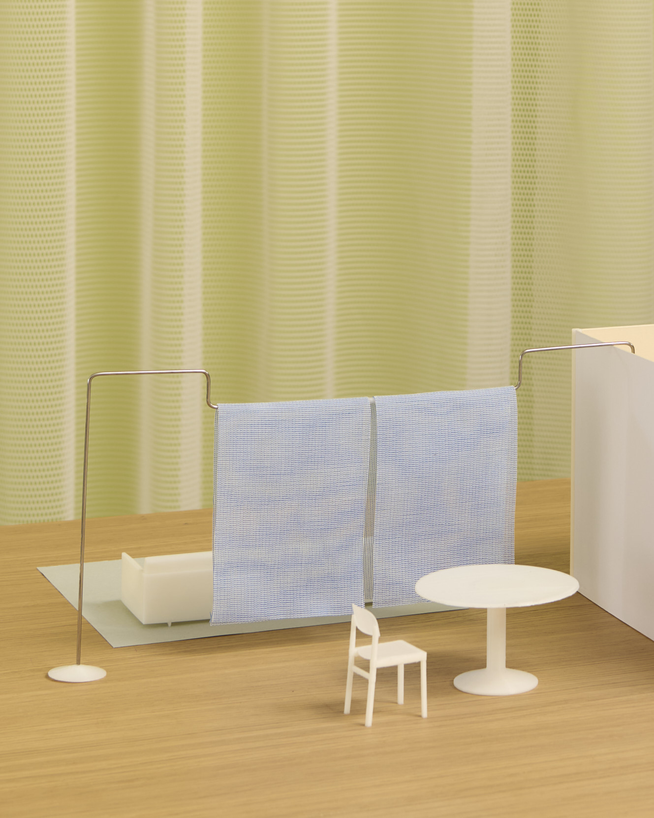 A miniature architectural model of the "Veil" system on a wooden tabletop. Two fine metal rods support sheer blue mesh panels, separating a small white lounge area from a round table and chair. A green mesh curtain in the background echoes the partition’s light and airy character.