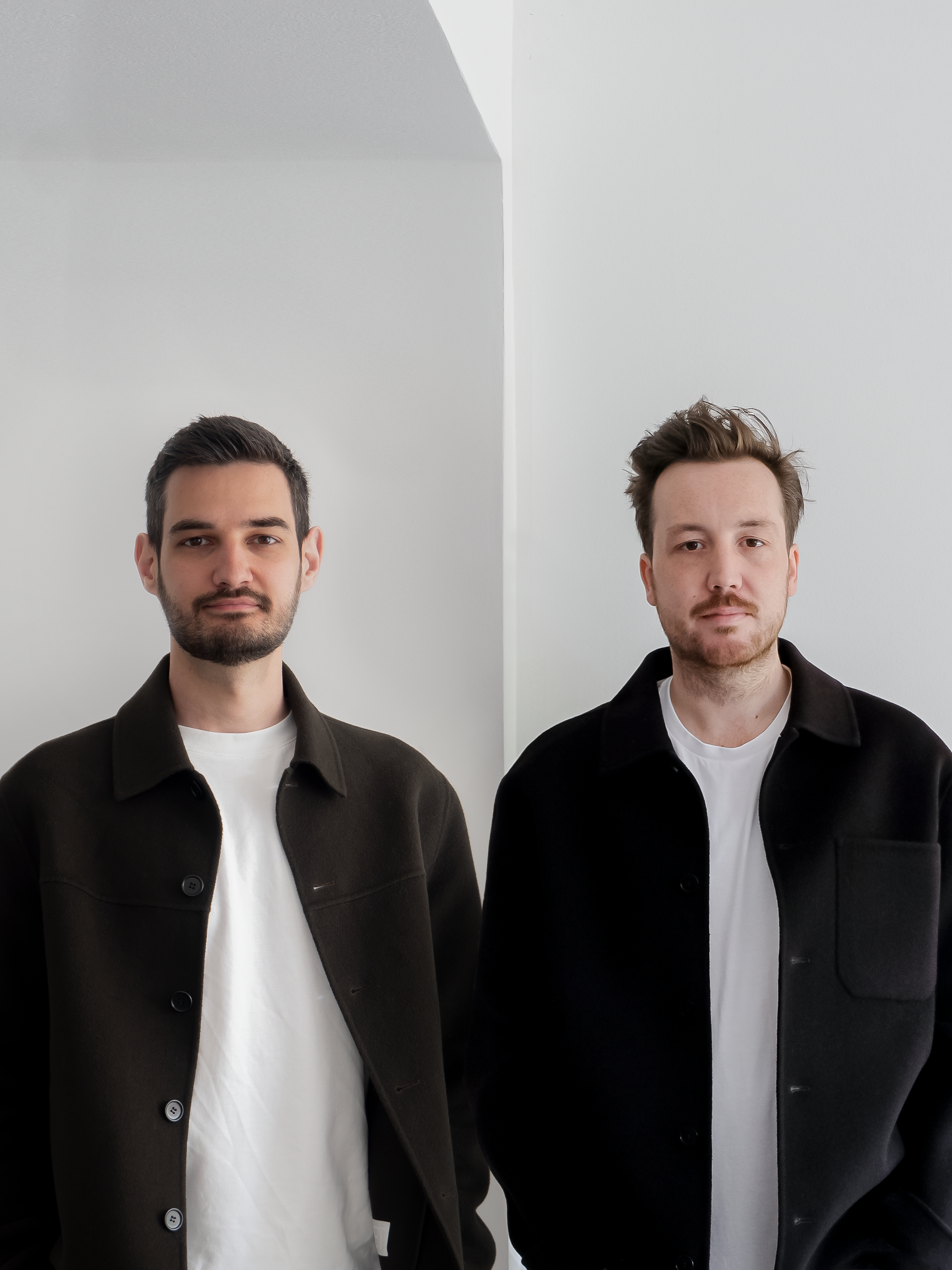 Portrait of designers Nikola & Florian standing side-by-side in a minimalist white interior. Both wear plain white T-shirts under dark buttoned jackets. 