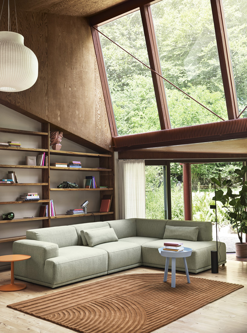A modern living room with large windows showcasing a lush green forest view. The room includes a light green Connect Soft Modular Sofa, a small blue side table, and a white ribbed pendant light, creating a serene reading corner.