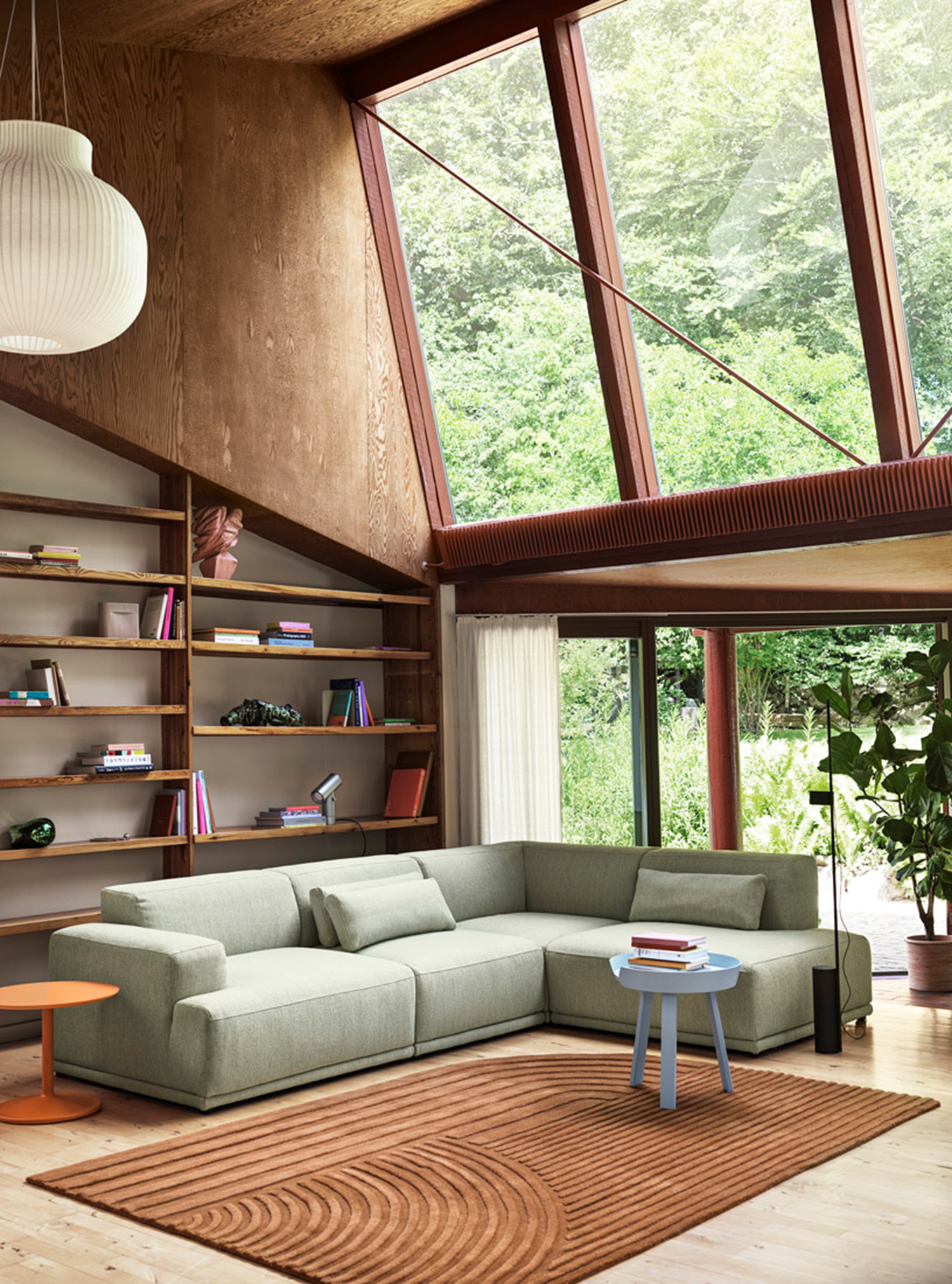 A modern living room with large windows showcasing a lush green forest view. The room includes a light green Connect Soft Modular Sofa, a small blue side table, and a white ribbed pendant light, creating a serene reading corner.