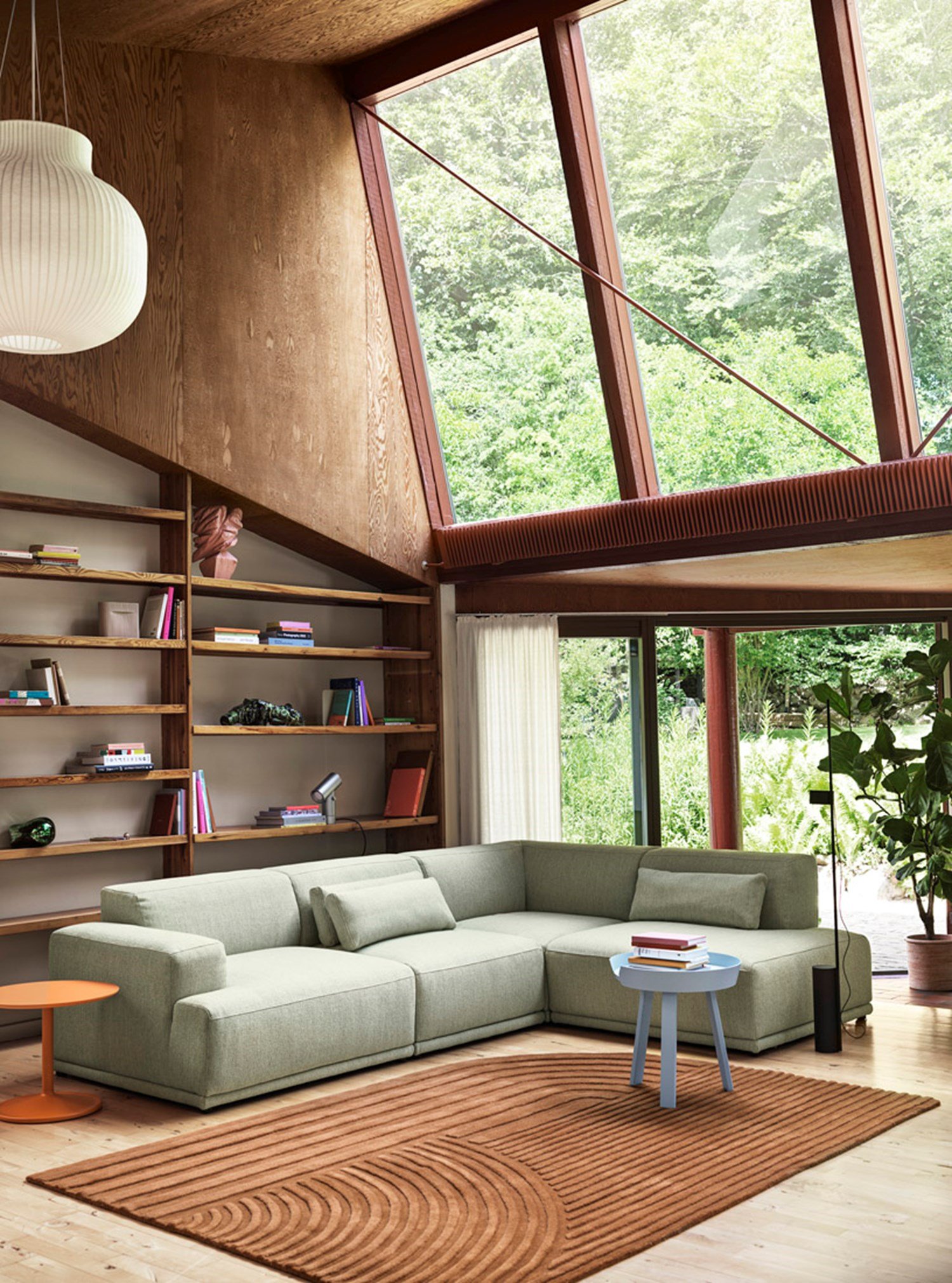 A modern living room with large windows showcasing a lush green forest view. The room includes a light green Connect Soft Modular Sofa, a small blue side table, and a white ribbed pendant light, creating a serene reading corner. A modern living room with large windows showcasing a lush green forest view. The room includes a light green Connect Soft Modular Sofa, a small blue side table, and a white ribbed pendant light, creating a serene reading corner.