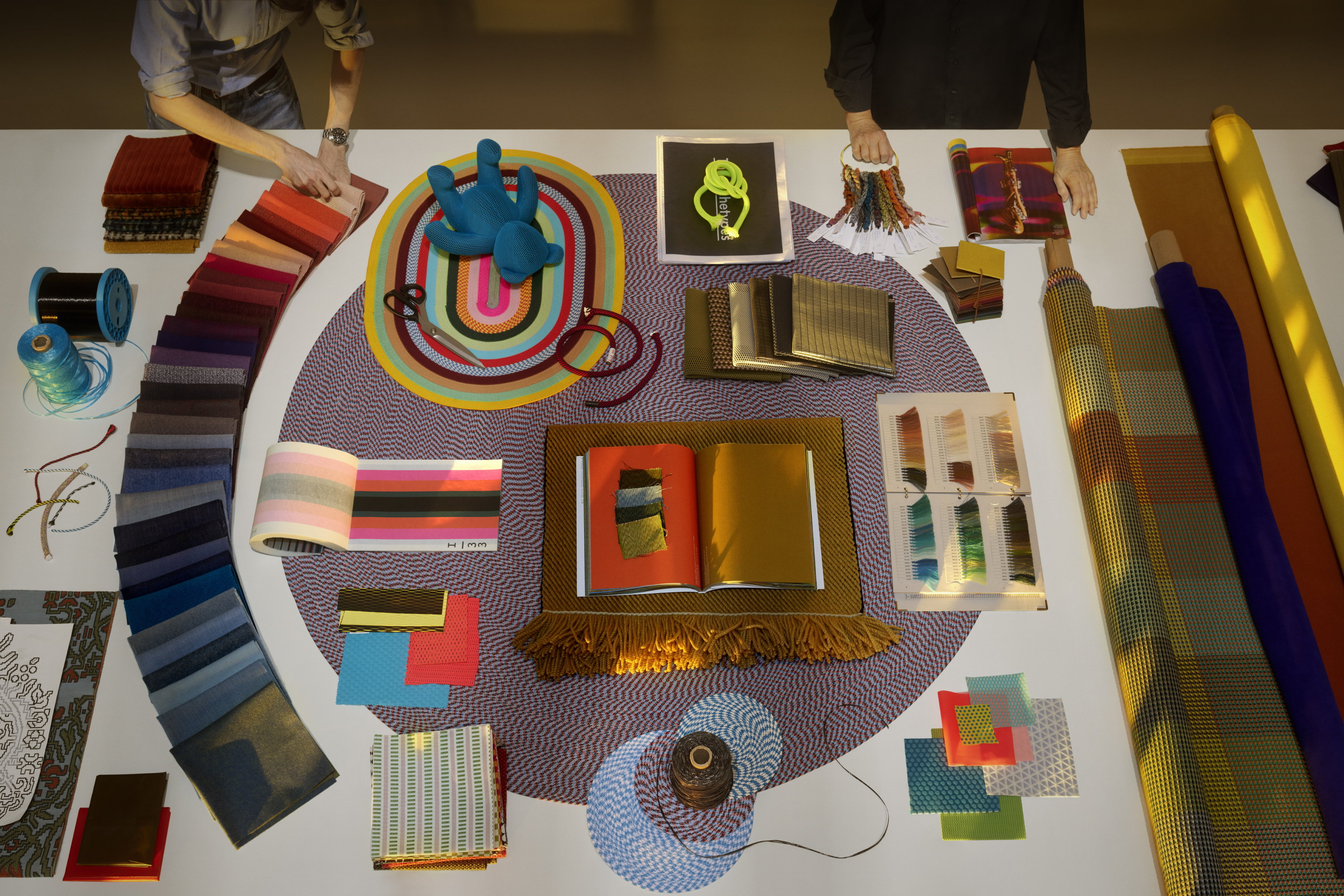Overhead view of a design studio table with scattered textile samples and creative tools, centered around a colorful arrangement.