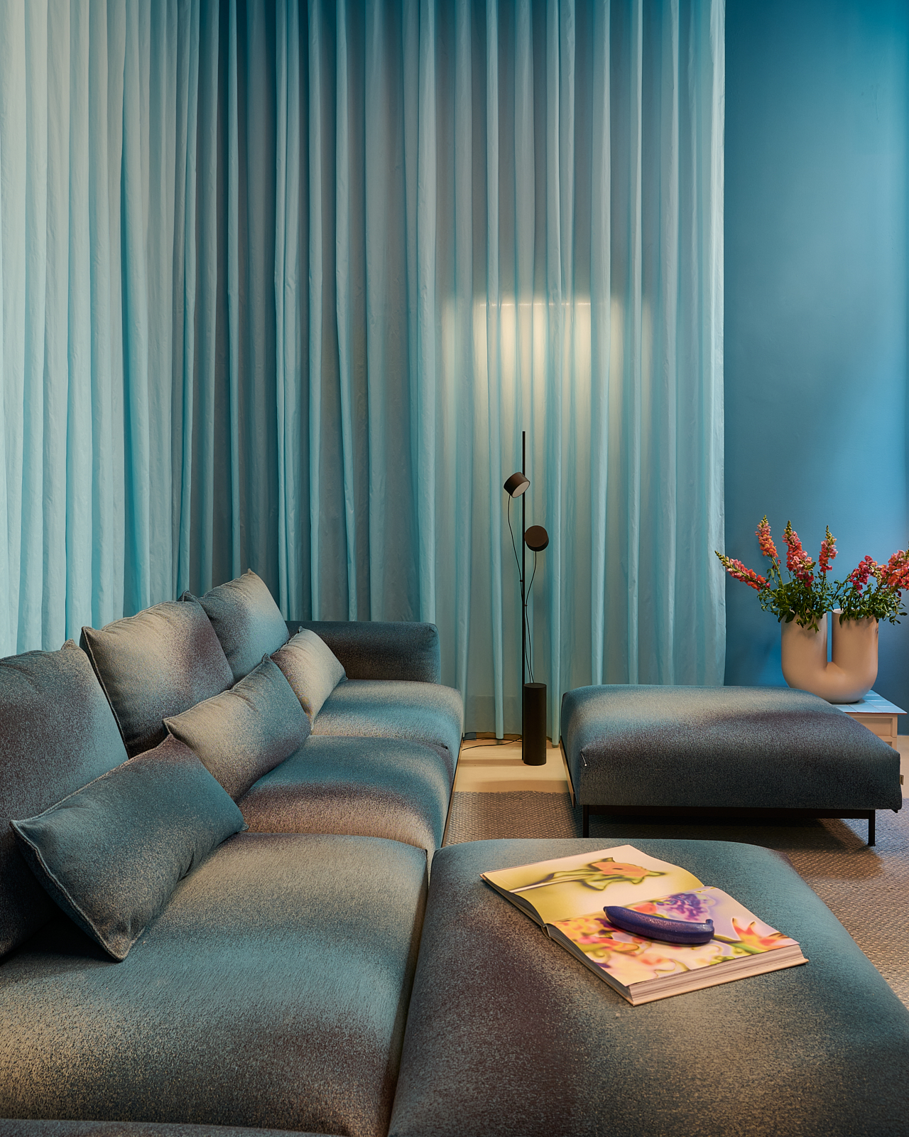 A blue room with blue curtains in Muuto's Milan apartment for Milan Design Week 2025. A big grey/blue sofa is placed in the room together with a table lamp and a big sculptural vase with red flowers. 