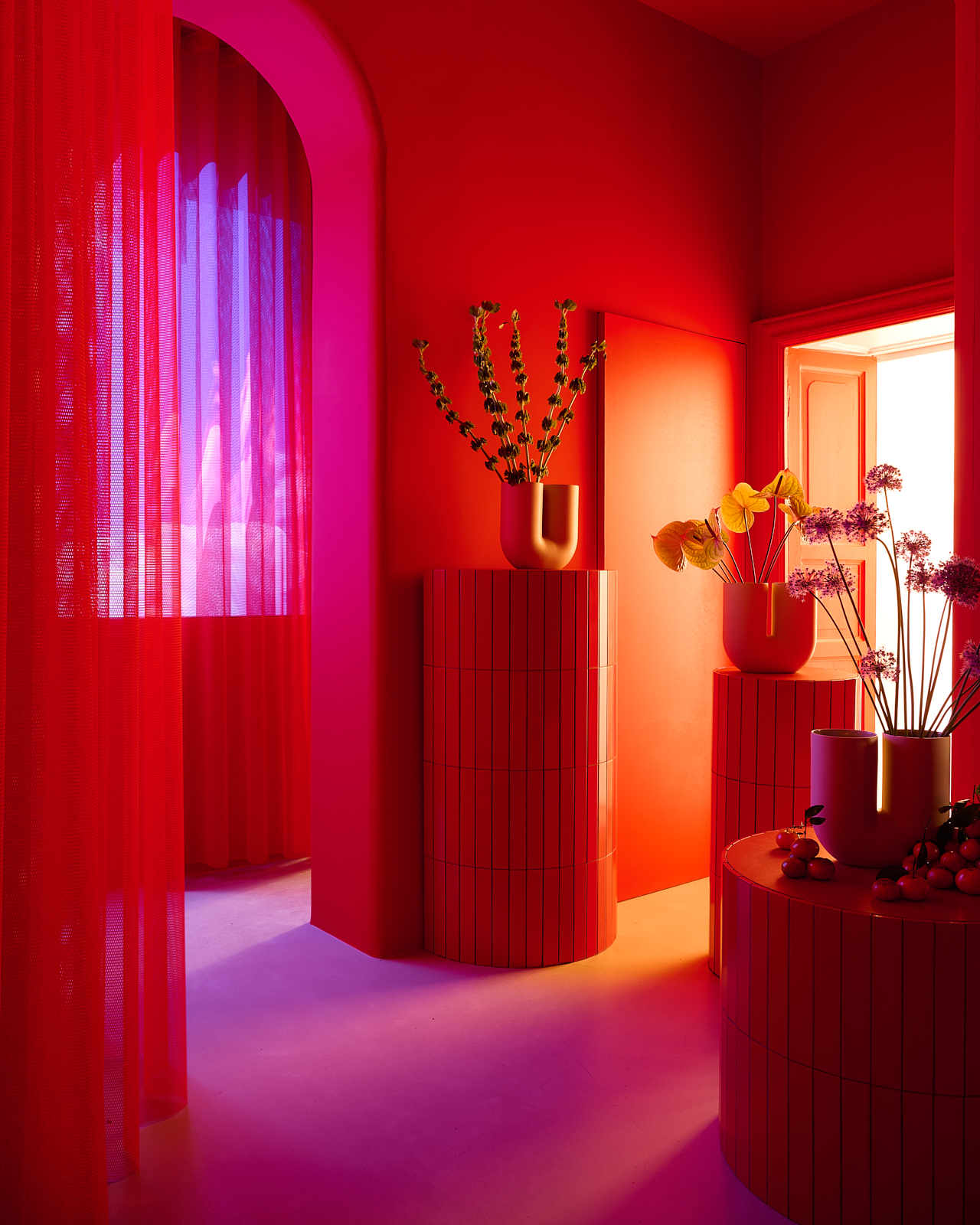 A red room with red curtains in Muuto's Milan apartment for Milan Design Week 2025. Sculptural vases with flowers are placed arround the room on stone pedistals in different heights. 