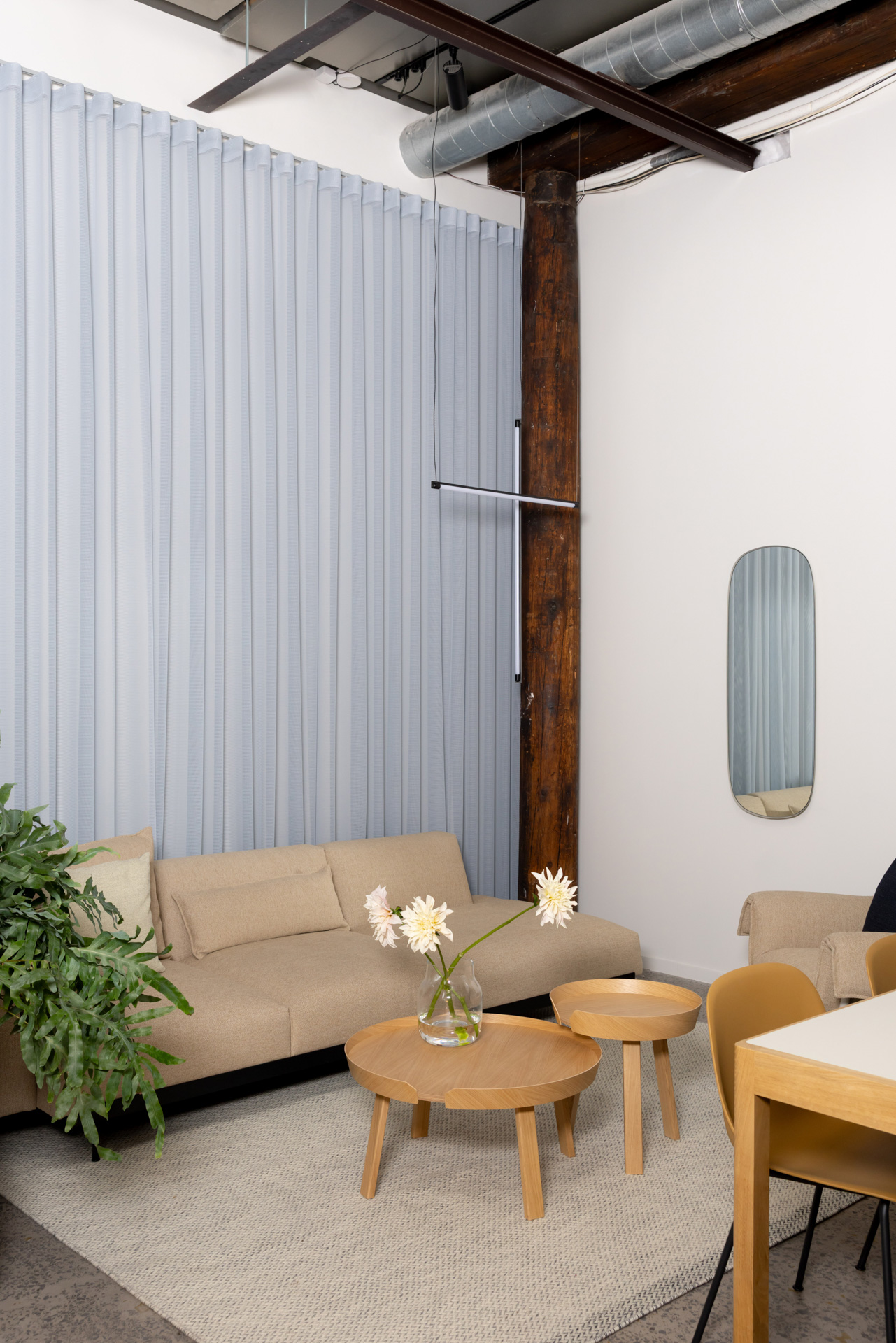 A sofa setting featuring a light beige sofa and a round wooden coffee table, placed on a sand-colored rug. An oval mirror hangs on the wall, while blue ceiling-high curtains behind the sofa create a cozy atmosphere.