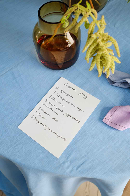 A handwritten recipe card with a burgundy glass vase and greenery on a blue tablecloth.
