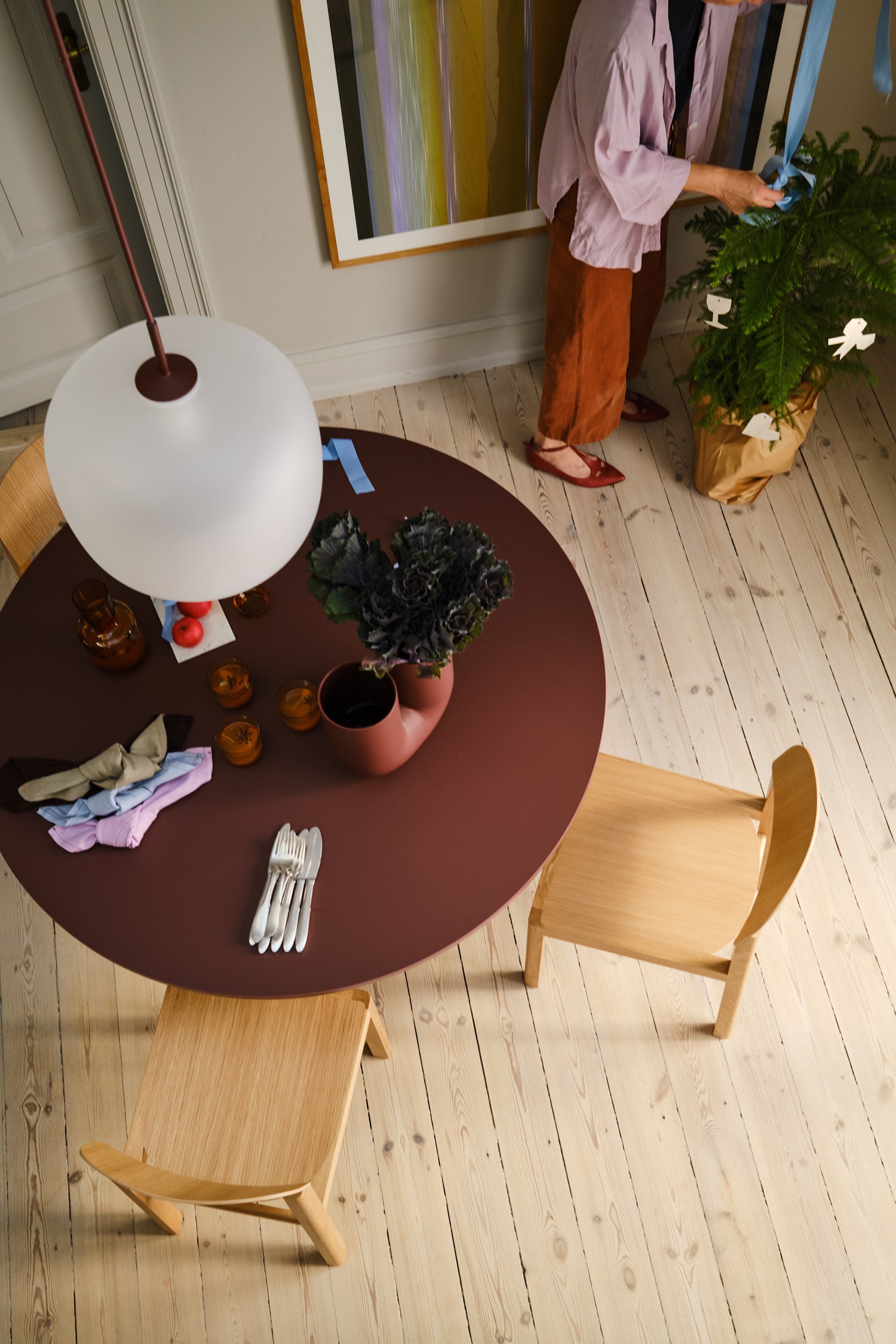 A woman in a lavender blouse arranges blue ribbons around a green potted plant. The table nearby features folded napkins, a deep red vase with kale, and a few glasses. A woman in a lavender blouse arranges blue ribbons around a green potted plant. The table nearby features folded napkins, a deep red vase with kale, and a few glasses.