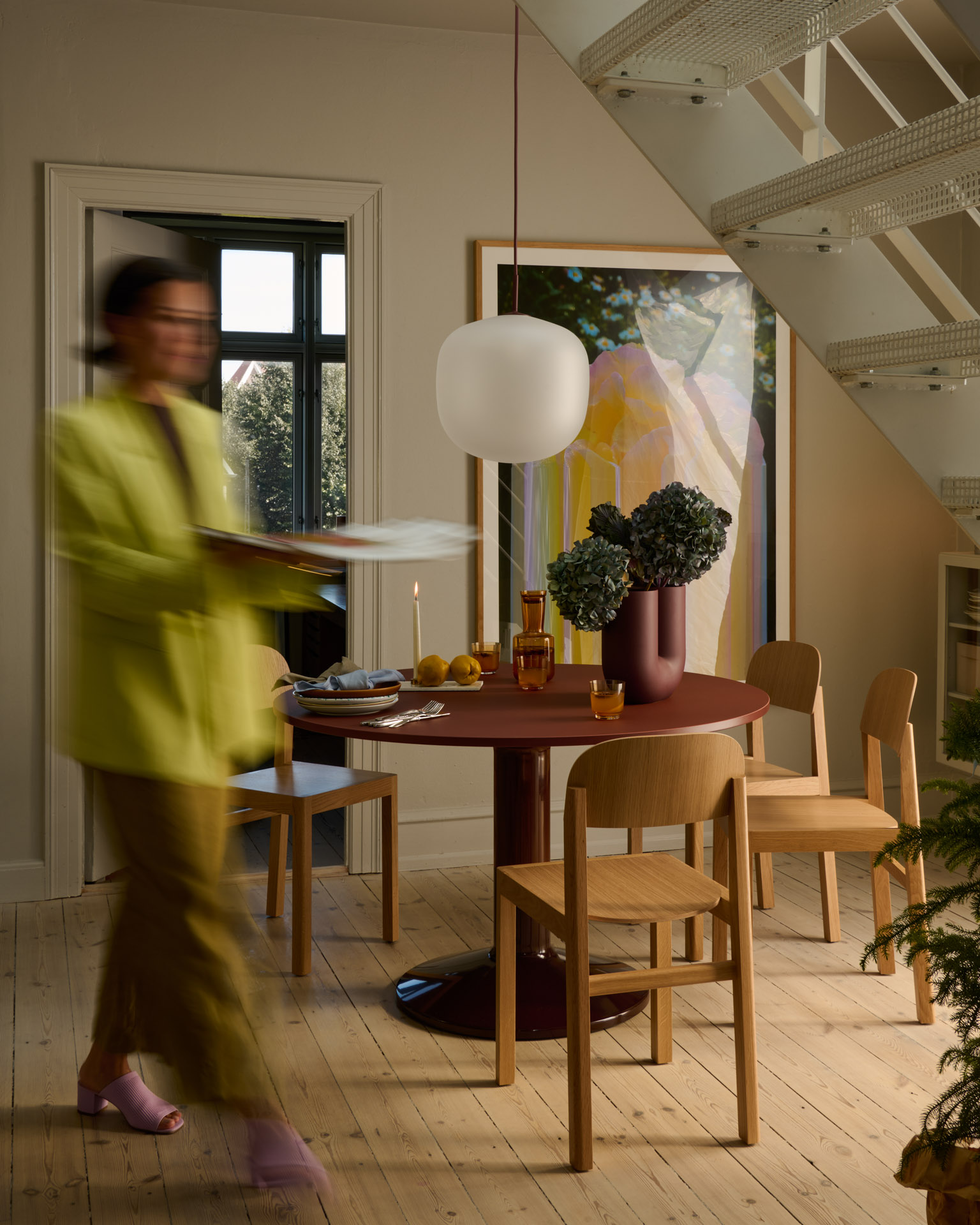 A woman walks past a round deep red table holding a vase with kale, two glasses, and plates. A soft light from a large window highlights the warm and cozy atmosphere, with a large artwork visible in the background.