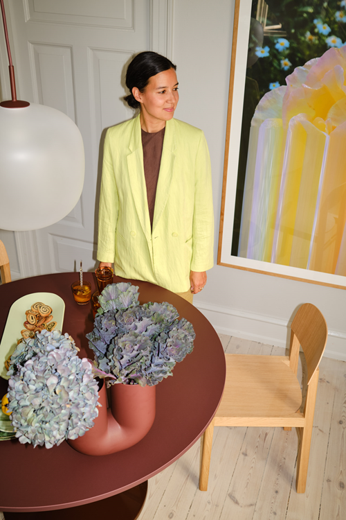 A woman wearing a lime green jacket stands next to a large artwork while smiling. The table in front of her holds purple kale arrangements and a tray of cookies. 