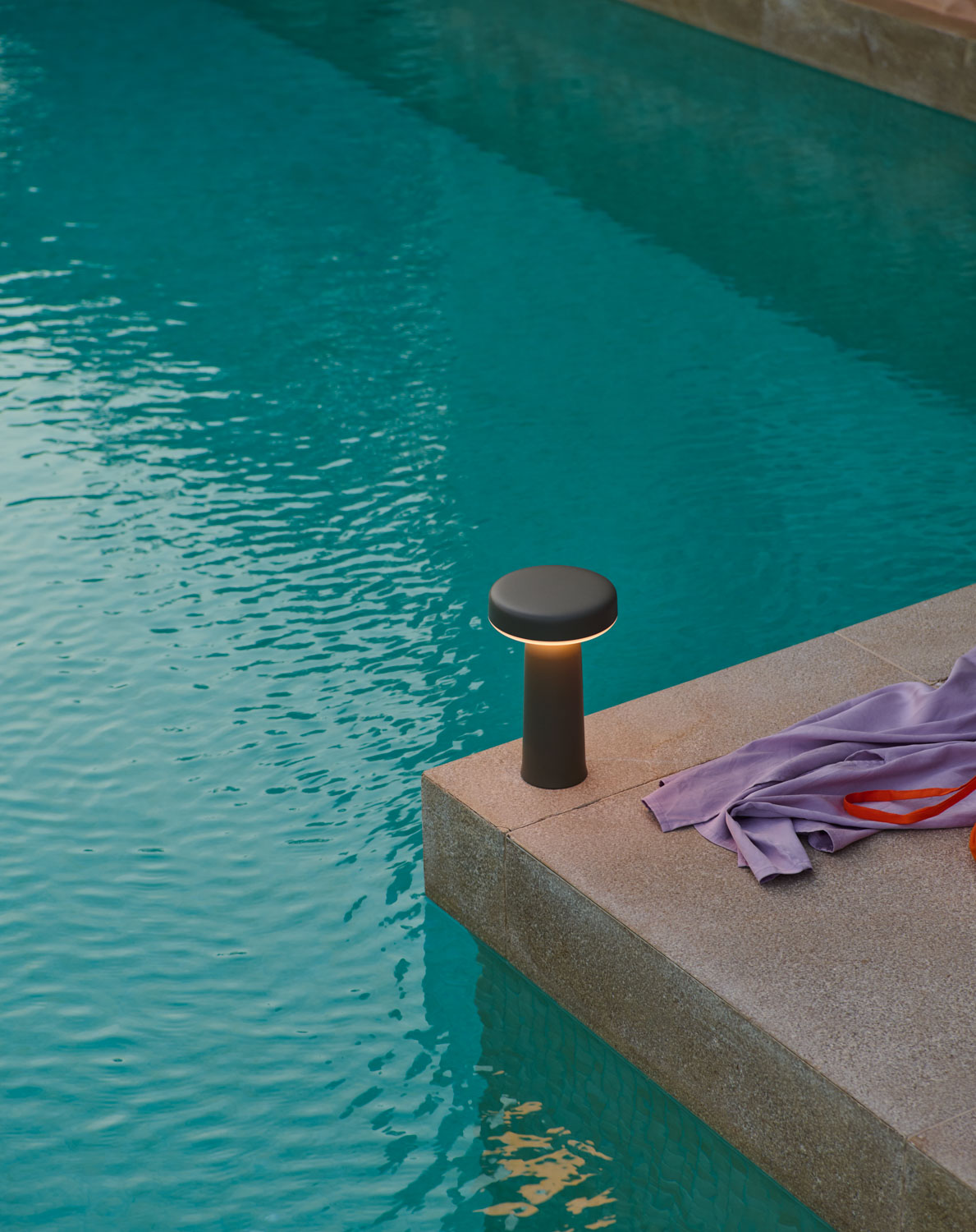 A sleek, modern Ease Lamp emits a warm glow on the edge of a swimming pool, next to a casually draped towel.
